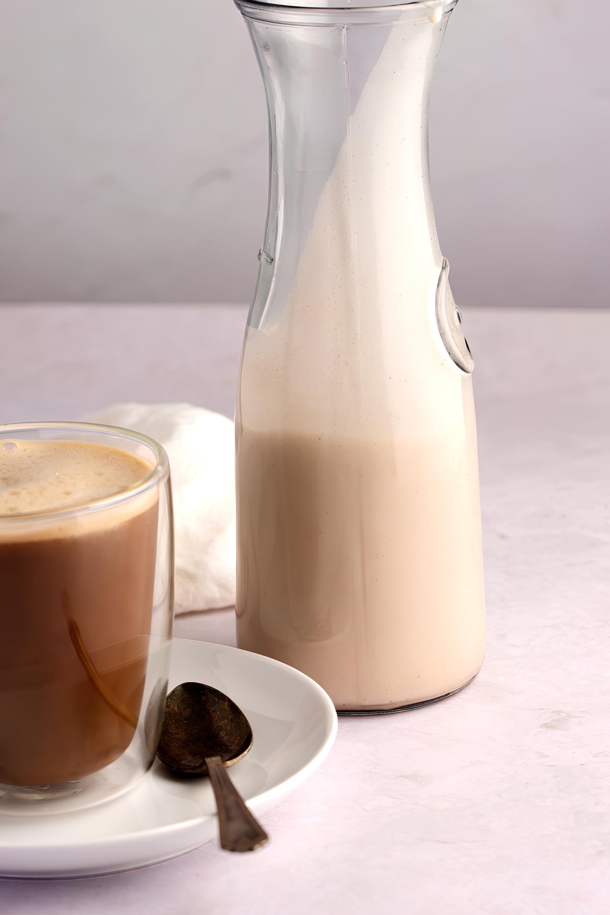 Long neck glass pitcher half filled with coconut creamer and a cup of foamy coffee in a glass mug. 